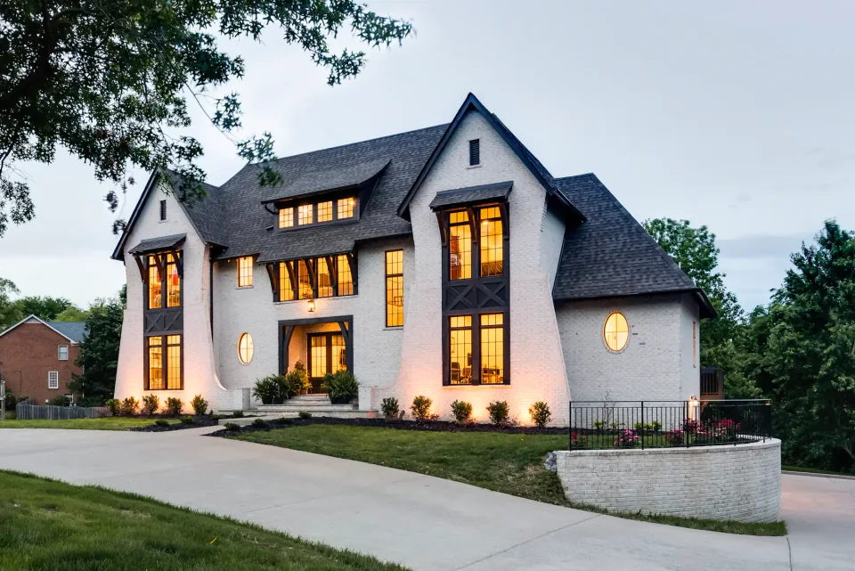White gabled home with warm window light beside a curved drive at dusk.