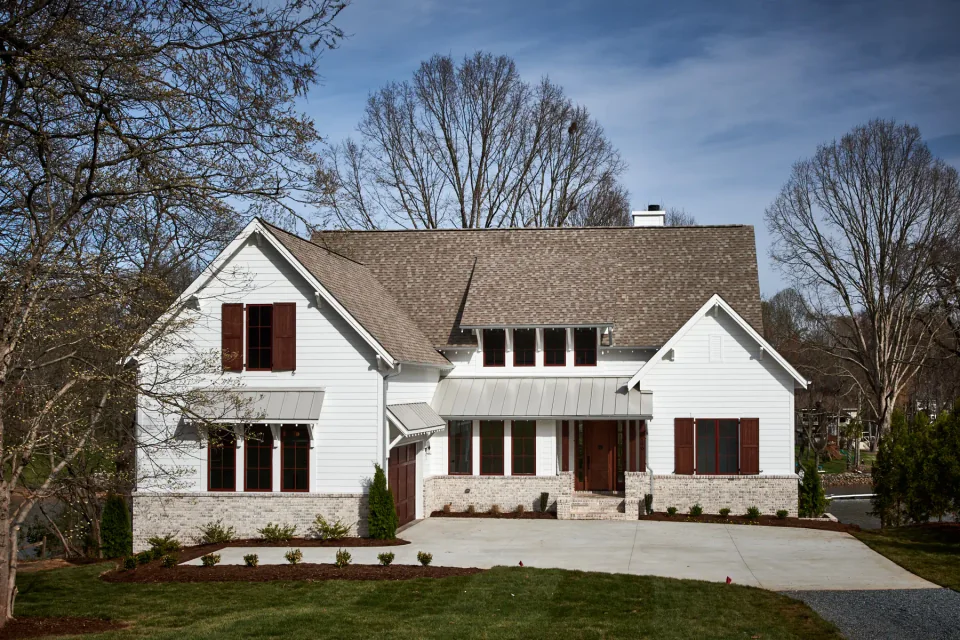White home with a covered porch, dark shutters and a wooded backdrop.