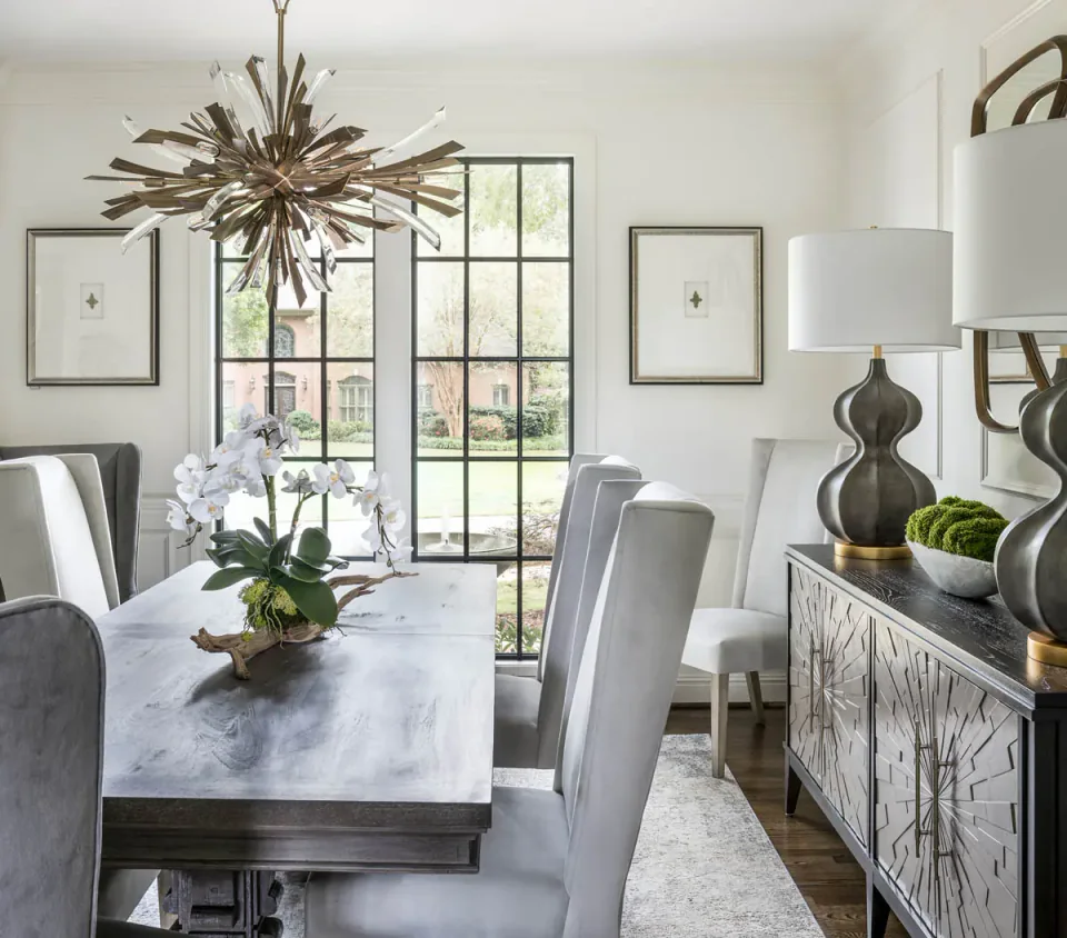 Dining room with a round table, upholstered chairs and large dark framed windows.