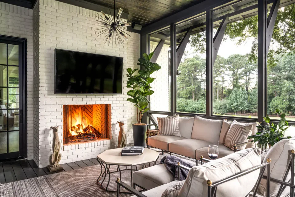 Screened porch with cushioned seating and a lit fireplace overlooking trees.